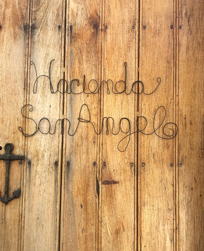 Gateway to accessibility, a copper wire sign that says Hacienda San Angel hanging on the wooden entry door of the hotel.
