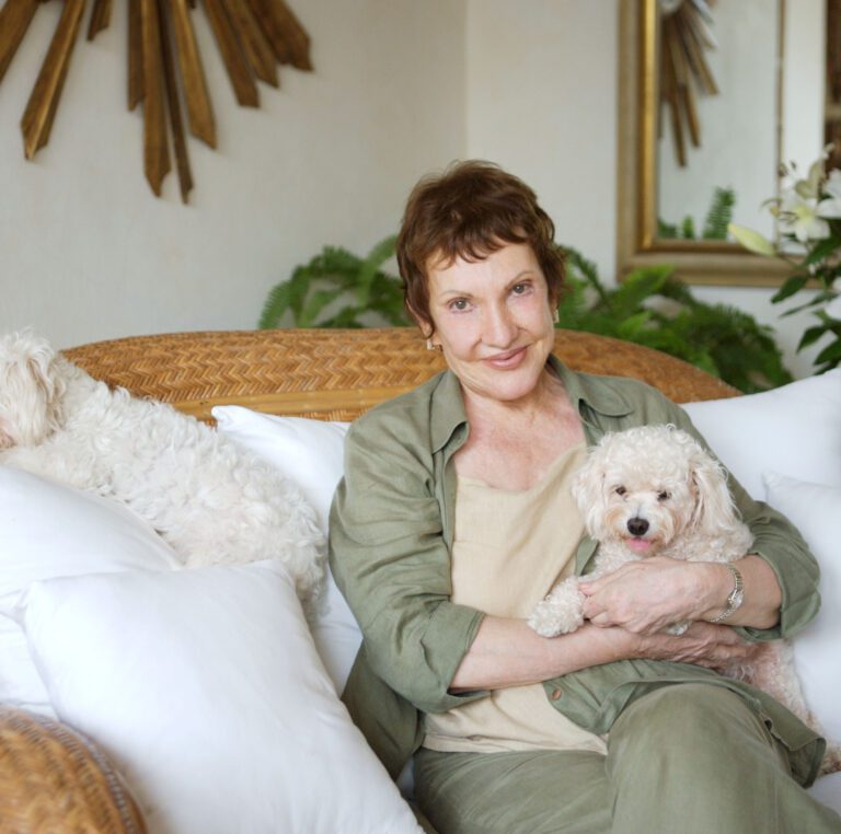 La Doña, Janice Chatterton, the founder of Hacienda San Angel is seated with her two small dogs on a wicker couch with white pillows surrounding them.