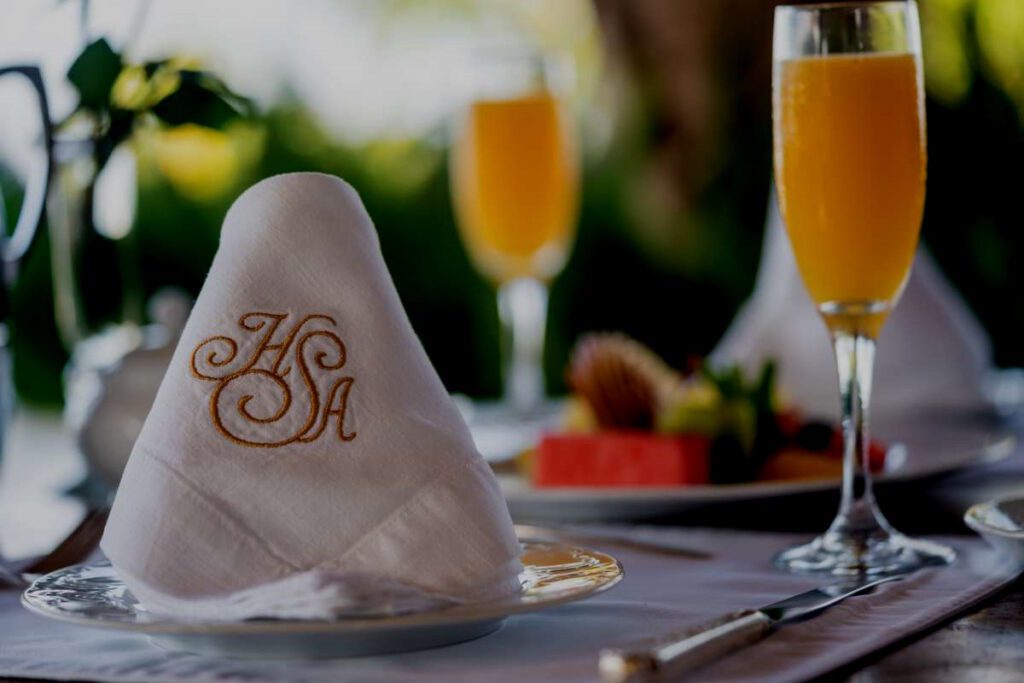 Elegant table arrangement featuring a flute of fresh orange juice, a white linen napkin with a gold embroidered HSA, and a plate of fresh fruit.