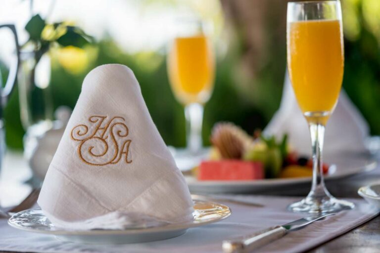 Elegant table arrangement featuring a flute of fresh orange juice, a white linen napkin with a gold embroidered HSA, and a plate of fresh fruit.