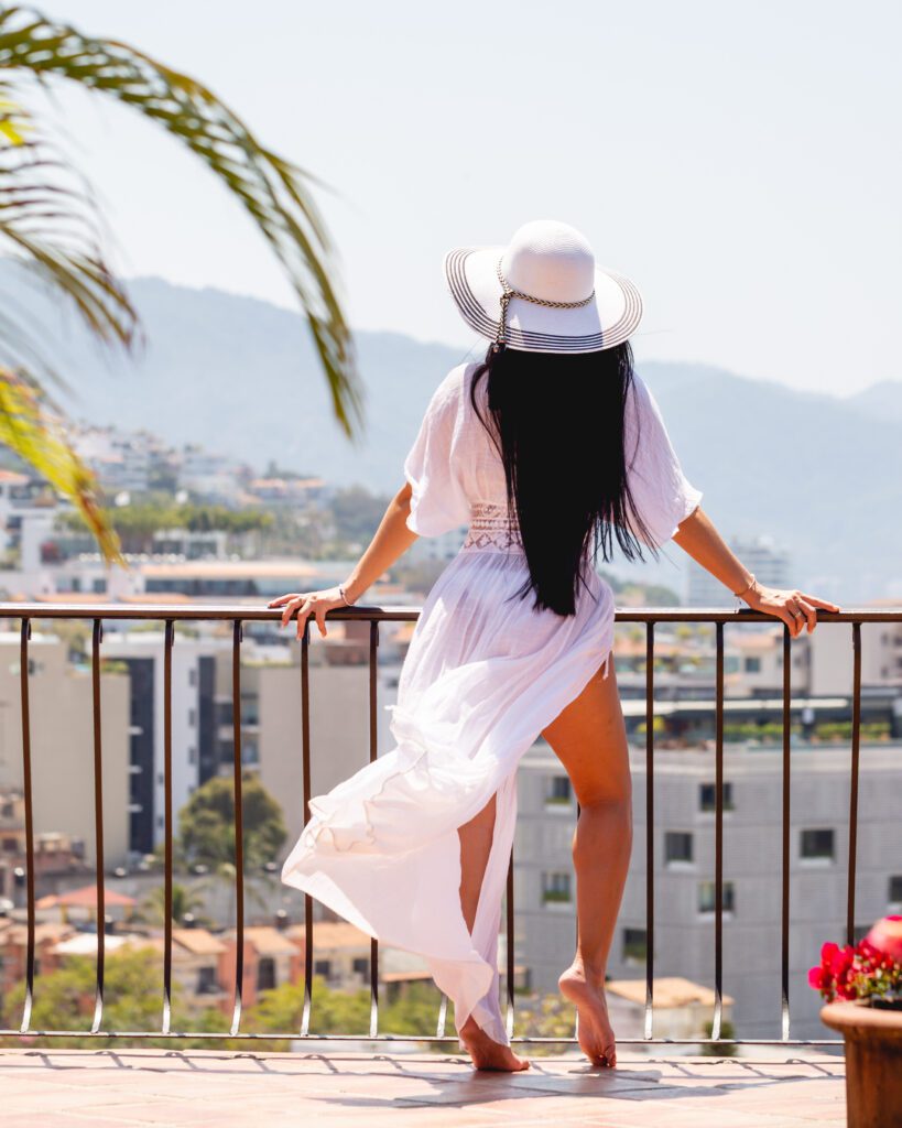 A woman with long black hair, wearing a white sun hat and a flowing white dress, standing on HSA's San Miguel terrace overlooking the Sierra Madre Mountains.