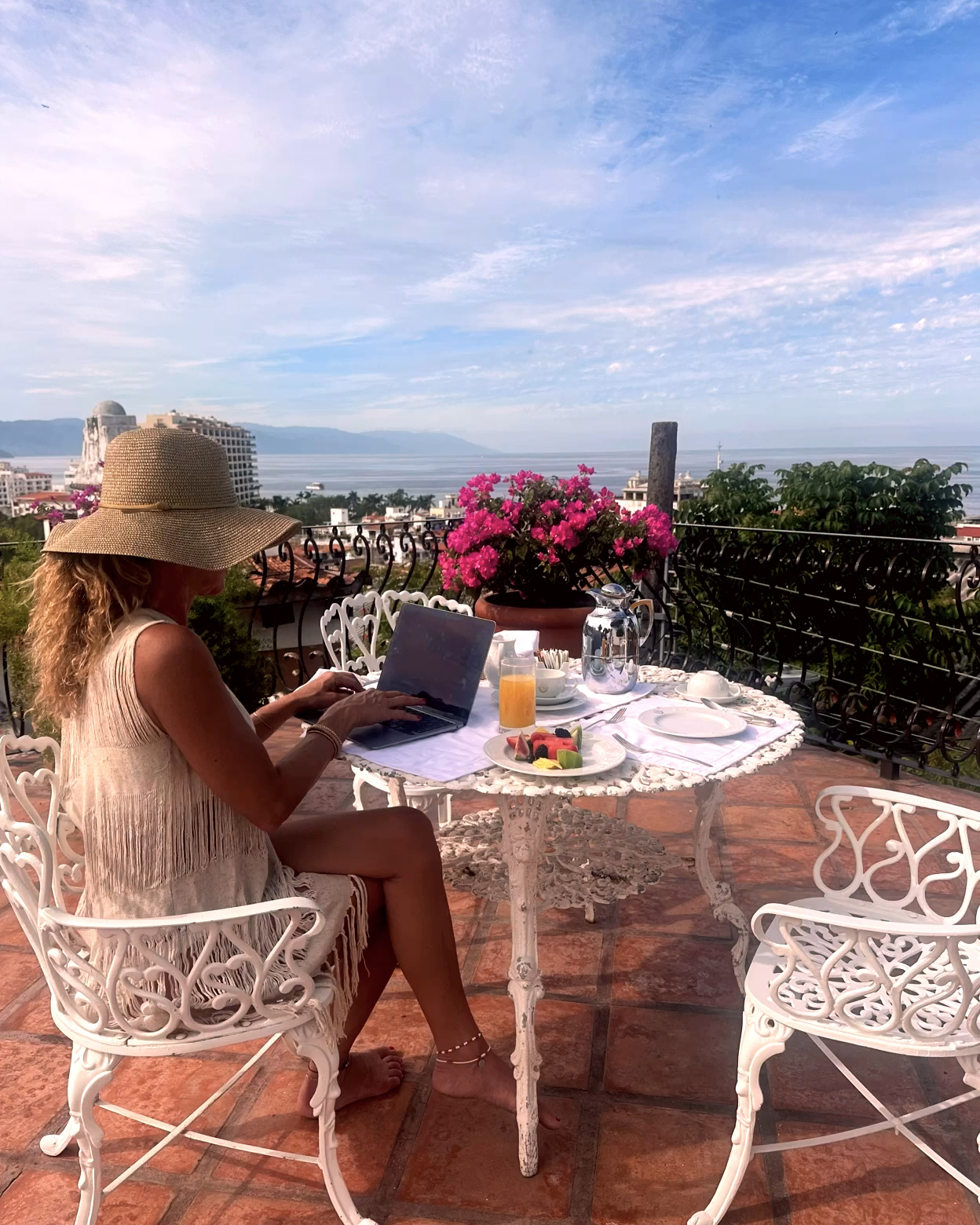 A woman sits on a terrace, enjoying breakfast while working on her laptop, surrounded by bougainvillea and overlooking Banderas Bay in the morning.