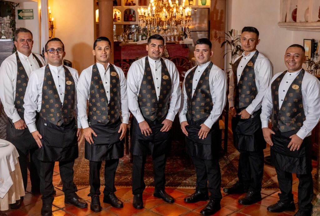 Reservation Policy showing seven waiters, ready to serve a welcome drink they are dressed in uniform, lined up in front of the restaurant's cantina under an elegant chandelier.