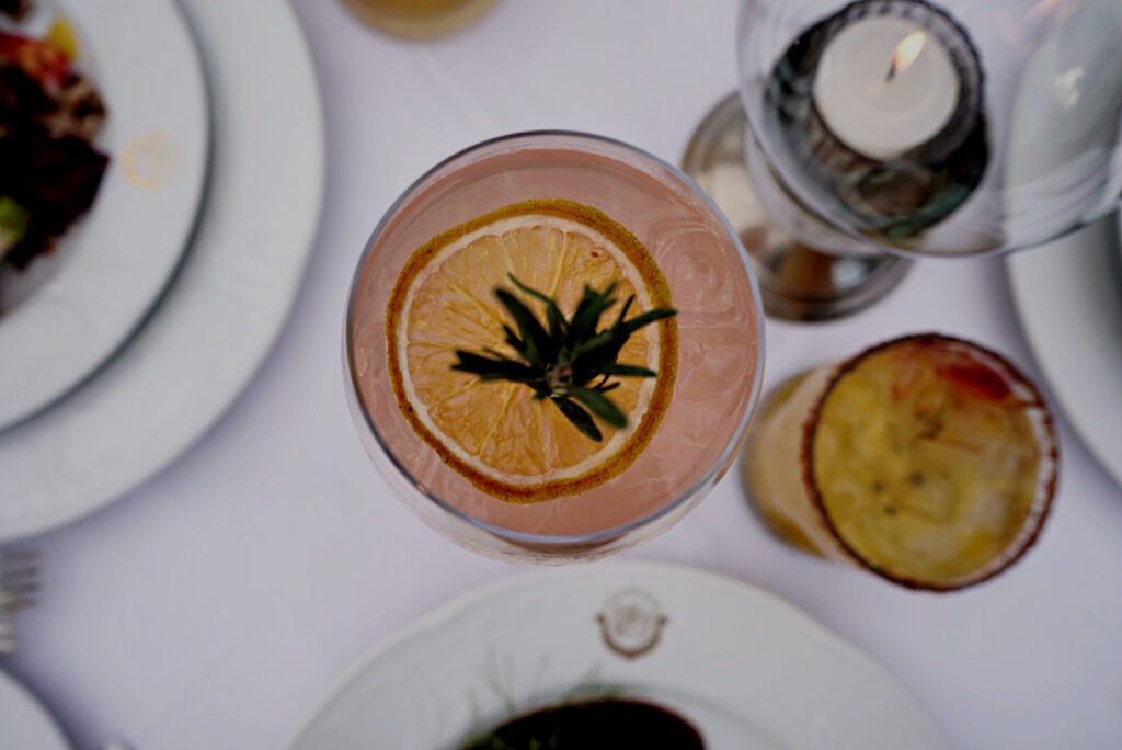 Exquisite table setting from a bird's-eye view showcasing a pink cocktail garnished with an orange slice and a sprig of rosemary.